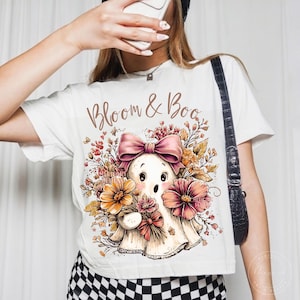 May include: A white t-shirt featuring a graphic of a ghost with a pink bow, surrounded by autumn-colored flowers and foliage. The text "Bloom & Boo" is printed in a brown script font above the ghost illustration. The shirt is paired with black and white checkered bottoms.