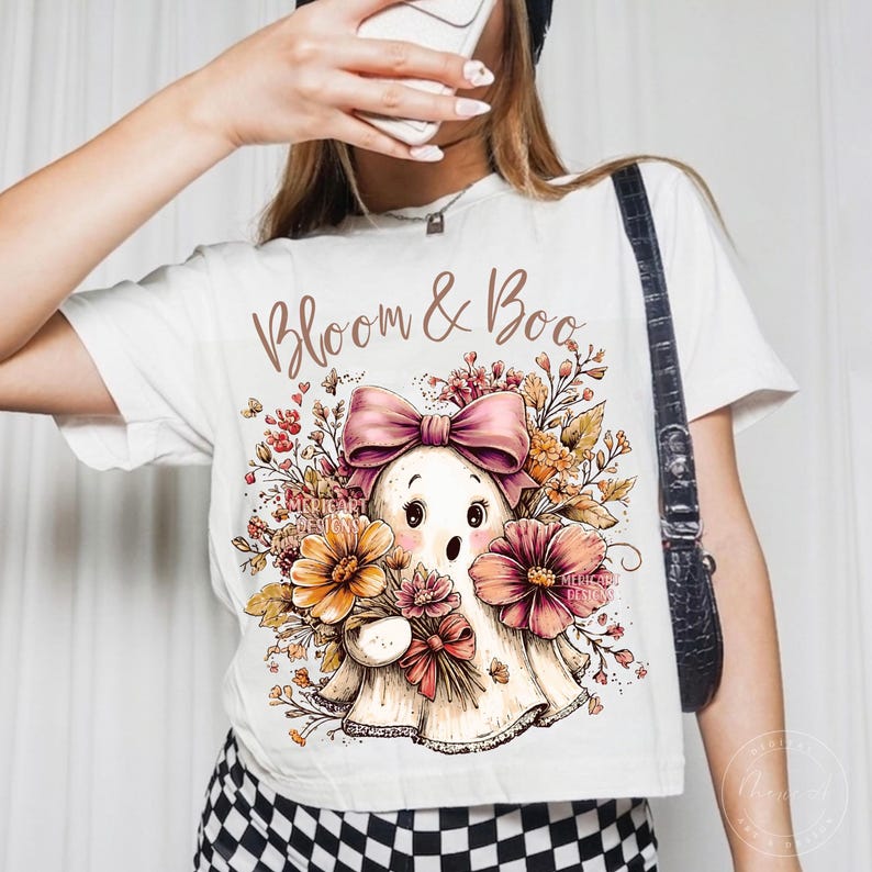 May include: A white t-shirt featuring a graphic of a ghost with a pink bow, surrounded by autumn-colored flowers and foliage. The text "Bloom & Boo" is printed in a brown script font above the ghost illustration. The shirt is paired with black and white checkered bottoms.
