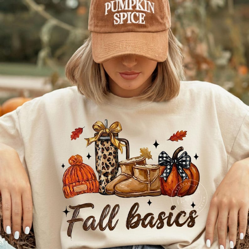 May include: A beige sweatshirt featuring a graphic of an orange knit beanie, a leopard print tumbler with a gold bow, tan boots, and an orange pumpkin with a black and white plaid bow. The text "Fall basics" is printed below the graphic.