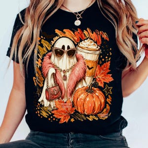Puede incluir: Una camiseta negra con el gr&aacute;fico de un fantasma con gafas de sol de ojo de gato, una estola de piel sint&eacute;tica rosa y un collar dorado, sosteniendo un bolso rojo. Junto al fantasma hay una calabaza y un caf&eacute; rodeados de hojas de oto&ntilde;o.