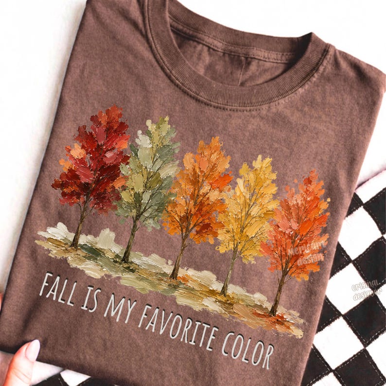 K&ouml;nnte beinhalten: Ein braunes T-Shirt mit Rundhalsausschnitt und einem Aufdruck von f&uuml;nf B&auml;umen mit strukturierten, malerischen Bl&auml;ttern in Rot-, Gr&uuml;n-, Orange- und Gelbt&ouml;nen. Unter den B&auml;umen steht der Text "FALL IS MY FAVORITE COLOR" in wei&szlig;er Schrift.