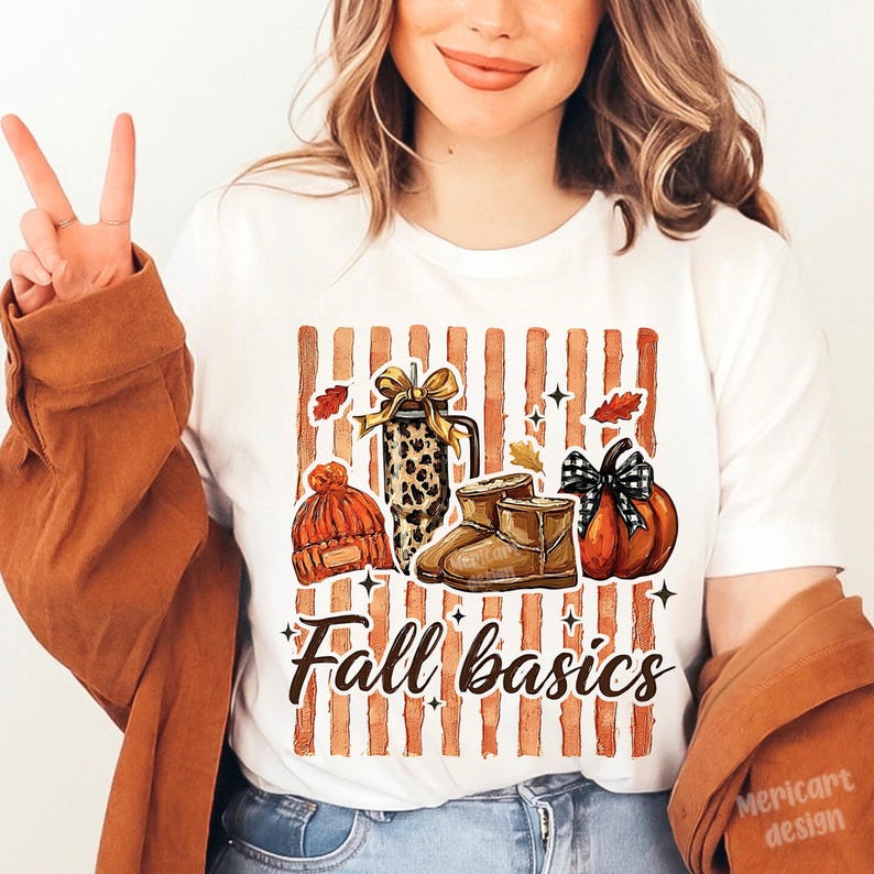 May include: A white t-shirt featuring a graphic print of fall items including an orange knit beanie, a leopard print tumbler with a gold bow, tan boots, and a pumpkin with a black and white plaid bow, over vertical stripes with the text Fall basics.