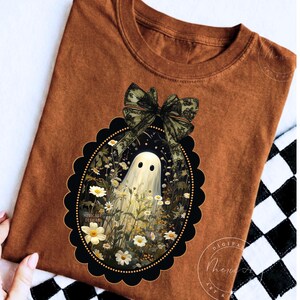 May include: A rust-colored t-shirt featuring a graphic of a white ghost standing in a field of white daisies, enclosed within a black scalloped oval frame topped with a large, detailed green lace bow. The text Mericade Designs is visible.