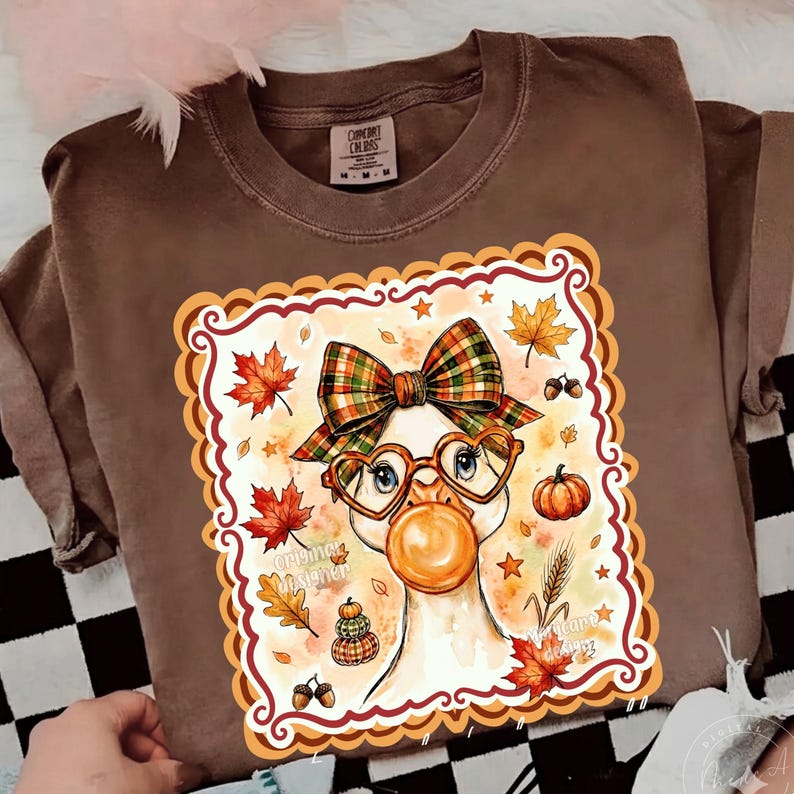 May include: A brown t-shirt featuring a graphic of a goose wearing a plaid bow and heart-shaped glasses while blowing a bubble. The design includes autumn leaves, pumpkins, and acorns, all framed within a decorative scalloped border.