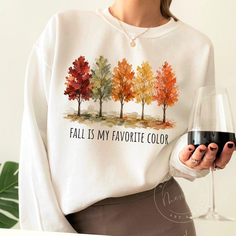 K&ouml;nnte beinhalten: Ein wei&szlig;es Sweatshirt mit Rundhalsausschnitt und einer Grafik von f&uuml;nf B&auml;umen in herbstlichen Farben wie Rot, Orange, Gelb und Gr&uuml;n. Unter den B&auml;umen steht der Text "FALL IS MY FAVORITE COLOR" in einer einfachen schwarzen Schrift.