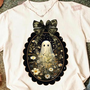 May include: A cream-colored t-shirt featuring a graphic of a white ghost standing in a field of white daisies and yellow wildflowers. The graphic is enclosed in a black scalloped oval frame topped with a large, sheer black lace bow.
