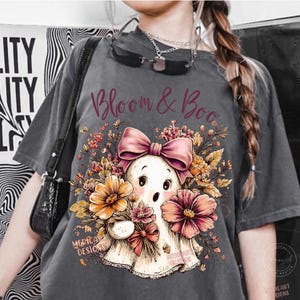 May include: A charcoal gray t-shirt featuring a graphic print of a white ghost wearing a large pink bow, surrounded by orange and pink flowers and foliage. The text "Bloom & Boo" is printed in a dark red script font above the ghost illustration.