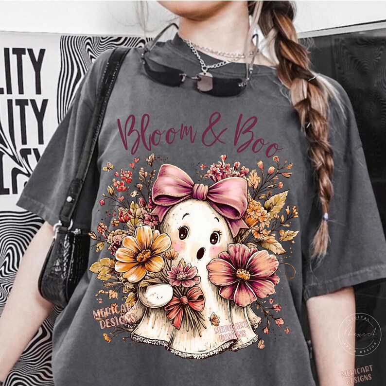 May include: A charcoal gray t-shirt featuring a graphic print of a white ghost wearing a large pink bow, surrounded by orange and pink flowers and foliage. The text "Bloom & Boo" is printed in a dark red script font above the ghost illustration.