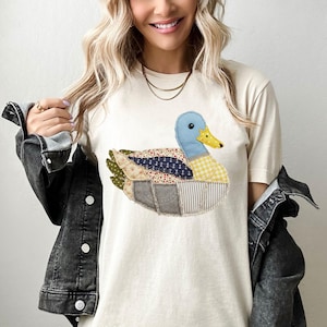 May include: Cream-colored t-shirt featuring a patchwork duck design. The duck has a blue head, yellow beak, and a body composed of various patterned fabrics, including floral, nautical, and gingham. The shirt is a casual, everyday wear item.
