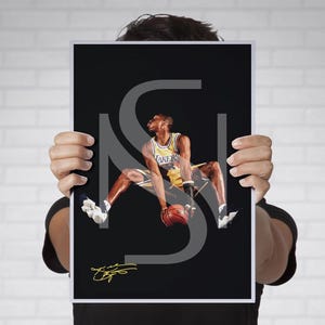 May include: A black and white photograph of a basketball player wearing a yellow and purple jersey with the word "LAKERS" on the front. The player is in mid-air, holding a basketball and about to dunk. The player's signature is in the bottom left corner of the photograph.
