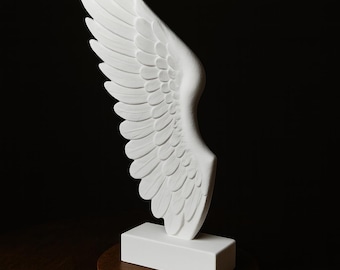 May include: A white, sculpted wing, likely a decorative item, is displayed against a dark background. The wing is mounted on a white rectangular base, which sits atop a round, dark wooden stand. The wing's feathers are intricately detailed.