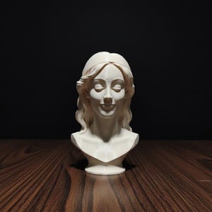 May include: A white bust sculpture of a woman with long, wavy hair and closed eyes. The sculpture is on a dark wooden surface with a black background. The bust is a classical art piece.