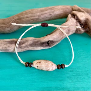 May include: A white string bracelet with a brown shell bead and brown wooden beads. The bracelet is on a piece of driftwood.