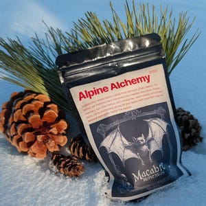 May include: A black pouch labeled "Alpine Alchemy" and "Macabre Mementos" sits in the snow next to pine needles and pine cones. The pouch features a bat illustration and text describing a winter blend.