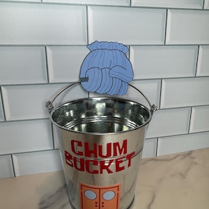 Puede incluir: Un cubo de metal galvanizado con asa plateada y las palabras "CHUM BUCKET" en letras rojas. Una imagen de dibujos animados azul está unida al asa. Debajo del texto hay un gráfico naranja de una puerta con dos ventanas redondas.