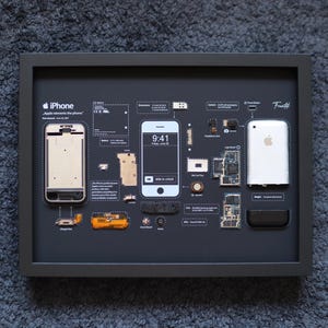 以下が含まれることがあります： iPhoneのコンポーネントを表示する額装されたアートワーク。黒い背景には、分解された電話、完全な電話、およびその他の部品があります。「iPhone」と「Apple reinvents the phone」というテキストが含まれています。