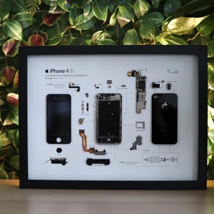 以下が含まれることがあります： iPhone 4S の分解図が描かれた額装されたプリント。デバイス内の個々のコンポーネントとその配置を示しています。プリントは白黒で、白い背景に黒いテキストが使用されています。テキストには「iPhone 4S」というタイトルとデバイスの説明が含まれています。