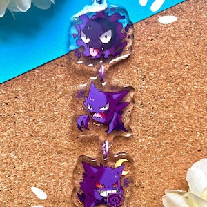 Puede incluir: Un llavero de Pokémon de tipo Fantasma morado y negro con tres versiones diferentes del personaje, Gengar. El llavero está hecho de acrílico transparente y tiene una cadena plateada.