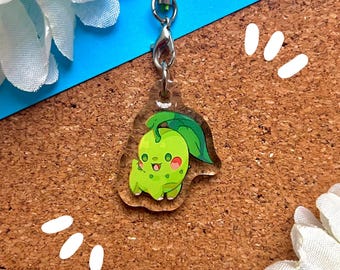 May include: A green and white cartoon character charm attached to a silver chain. The charm is shaped like a small, round creature with a leaf on its head. The charm is hanging from a clear acrylic keychain.