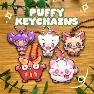 Puede incluir: Cinco llaveros acolchados con diseños de criaturas de dibujos animados en colores morado, blanco, marrón, rojo y rosa. Cada llavero está unido a una cadena de bolas de colores. El texto PUFFY KEYCHAINS aparece en letras verdes grandes.