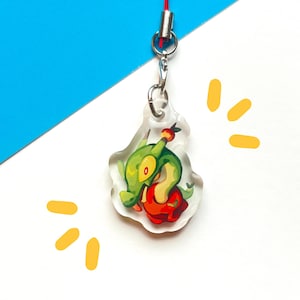Puede incluir: Un charm de acrílico transparente con un personaje de dibujos animados verde con un cuerpo rojo y naranja. El charm está sujeto a un cordón rojo y herrajes plateados. El fondo es blanco y azul con detalles amarillos.
