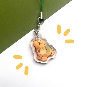 Puede incluir: Un dije de acrílico transparente con una criatura de dibujos animados de pelaje naranja claro, una mancha naranja más oscura en la espalda y un solo ojo grande. El dije está unido a un broche de langosta de metal plateado y un cordón verde fino.