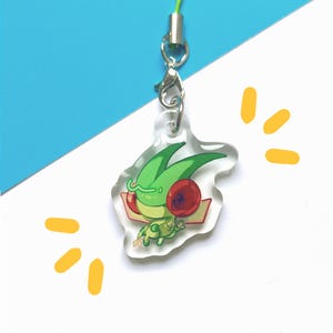 Puede incluir: Un charm de acrílico transparente con un personaje de dibujos animados verde con detalles rojos y hojas verdes. Está unido a un cordón verde y un cierre plateado. El fondo es blanco y azul con elementos gráficos amarillos.