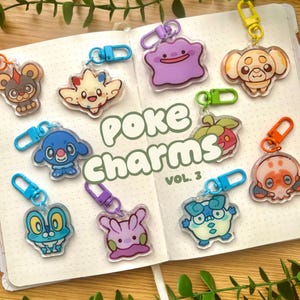 Pode incluir: Uma coleção de dez chaveiros de acrílico com vários designs de criaturas de desenhos animados, cada um preso a um fecho lagosta de plástico colorido. O texto "poke Charms VOL. 3" está impresso no centro da página do caderno aberto.