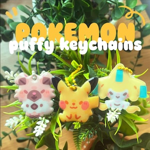 Può includere: Tre portachiavi Pokemon gonfi colorati. I portachiavi presentano una creatura rosa e bianca, un Pikachu giallo e un personaggio giallo e blu. I portachiavi sono appesi a catene gialle e sono circondati da fogliame verde. Il testo "Pokemon puffy keychains" è in alto.