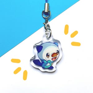 Puede incluir: Un charm de acrílico transparente con un personaje de dibujos animados de una nutria con detalles azules, blancos y marrones. El charm está sujeto a un cordón azul con un cierre plateado. El fondo es blanco y azul.