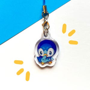 Puede incluir: Un charm de acrílico transparente con un personaje de dibujos animados de pingüino azul con vientre blanco y pies amarillos. El charm está unido a un cierre plateado y un cordón azul. El fondo es blanco y azul con detalles amarillos.