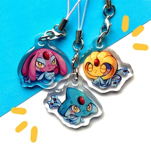 May include: Three clear acrylic charms featuring cartoon illustrations of Pokemon characters. The charms are pink, yellow, and blue with black outlines. Each charm has a silver metal ring attached to a string.