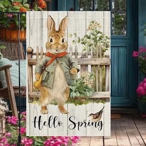 May include: A garden flag featuring a charming illustration of a rabbit dressed in a jacket and scarf, standing near a wooden fence. The flag has the words "Hello Spring" printed on it. The background includes a blue door and vibrant flowers.