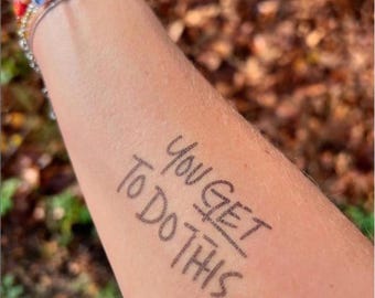 May include: Close-up of an arm with the handwritten phrase "You Get To Do This" in black ink. A beaded bracelet with the word "FEARLESS" is visible on the wrist, along with other bracelets. The background is blurred with fall foliage.