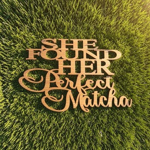 May include: A laser-cut wooden sign featuring the text "SHE FOUND HER" in a bold, block-style font, followed by the word "Perfect" and "Matcha" in a flowing, cursive script. The natural brown wood grain is visible throughout the lettering.