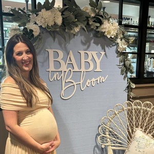 May include: A pregnant woman in a light beige dress stands in front of a blue backdrop with a white floral garland and a wooden sign that reads "Baby in Bloom". A white wicker chair with a floral patterned cushion sits to the right of the sign. The text "...was perfect" is written below the image.