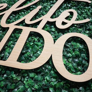 May include: Wooden sign with the words "Hello World" in a cursive font. The sign is made of light-colored wood and is set against a backdrop of green foliage. The letters are cut out, creating a three-dimensional effect.