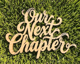 May include: A light brown wooden cutout sign featuring the words Our Next Chapter in a cursive script font. The sign has a smooth, flat surface and is laser-cut with intricate, flowing lettering that connects the words into a single piece.