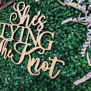 Puede incluir: Letrero de madera con la frase "She's Tying The Knot" en una elegante escritura. El letrero es de madera clara y est&aacute; sobre un fondo verde con cintas rosas e iridiscentes, ideal para una despedida de soltera o boda.