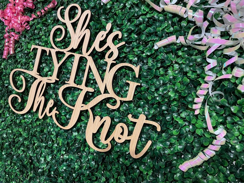 Puede incluir: Letrero de madera con la frase "She's Tying The Knot" en una elegante escritura. El letrero es de madera clara y est&aacute; sobre un fondo verde con cintas rosas e iridiscentes, ideal para una despedida de soltera o boda.