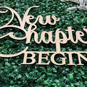 May include: Wooden sign with the words "New Chapter Begins" in a cursive font. The sign is light brown and sits on a bed of green foliage. Pink and white decorative accents are in the background.