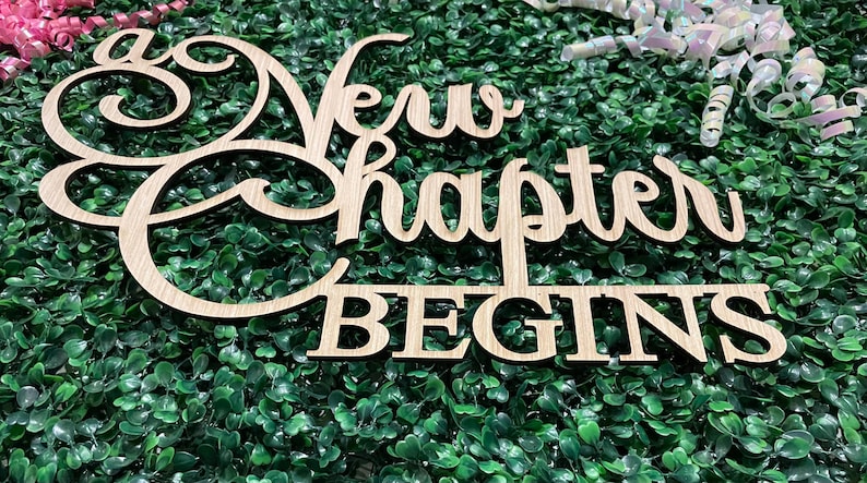 May include: Wooden sign with the words "New Chapter Begins" in a cursive font. The sign is light brown and sits on a bed of green foliage. Pink and white decorative accents are in the background.