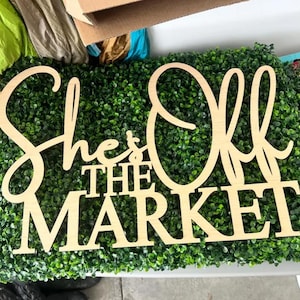 May include: A light-colored wooden sign with cut-out lettering that reads She's Off THE MARKET in a combination of cursive and block fonts, positioned against a dense, textured green artificial boxwood hedge backdrop.