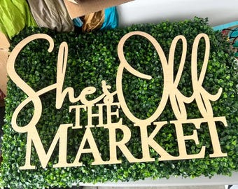 May include: A light-colored wooden sign with cut-out lettering that reads She's Off THE MARKET in a combination of cursive and block fonts, positioned against a dense, textured green artificial boxwood hedge backdrop.