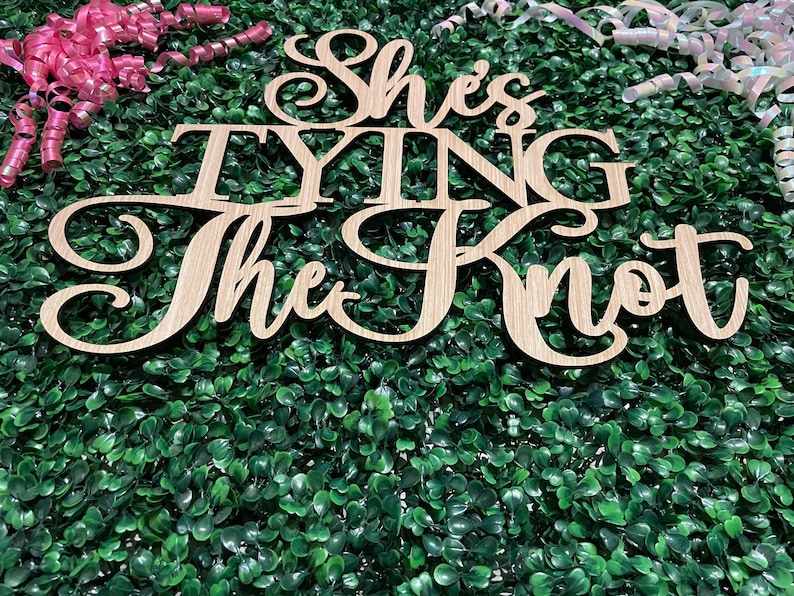 Puede incluir: Un letrero de madera con la frase "She's Tying The Knot" en fuente cursiva, sobre un fondo de follaje verde. Cintas rizadas rosas y blancas acent&uacute;an la escena, sugiriendo un evento festivo.