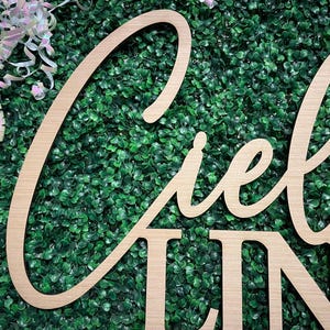 May include: Wooden sign with the word "Ciel" in a cursive font and the word "Line" in a block font. The sign is made of light-colored wood and is set against a backdrop of green foliage. White and pink ribbons are in the upper left corner.