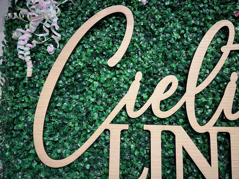 May include: Wooden sign with the word "Ciel" in a cursive font and the word "Line" in a block font. The sign is made of light-colored wood and is set against a backdrop of green foliage. White and pink ribbons are in the upper left corner.