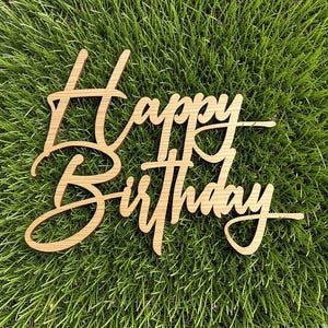 May include: A light brown wooden cake topper featuring the words Happy Birthday in a cursive script font. The letters are connected in a single piece, showcasing a natural wood grain texture throughout the surface of the cut-out lettering.