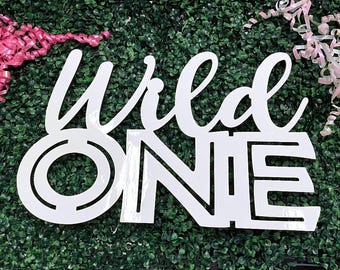 May include: A white sign featuring the text Wild ONE in a combination of cursive and block lettering. The word Wild is written in a flowing script font, while the word ONE is displayed in a bold, geometric sans-serif typeface with cutouts.