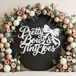 May include: A circular black wooden backdrop featuring the white 3D text Pretty Bows & Tiny Toes with a bow graphic. The sign is framed by an arch of balloons in shades of sage green, blush pink, and cream, interspersed with green foliage and roses.